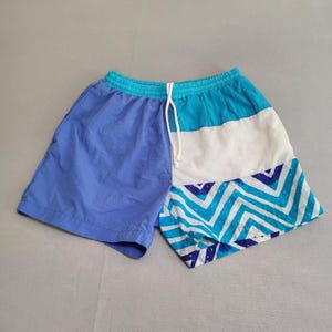 May include: Color-blocked swim trunks in shades of blue, white, and turquoise. The shorts feature a drawstring waistband and a geometric chevron pattern on the lower portion. Casual summer wear.