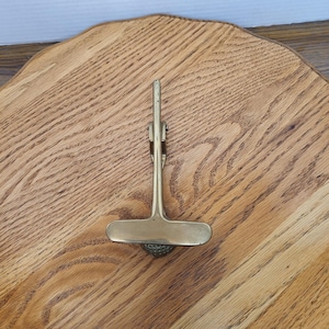 May include: A brass-colored golf club divot repair tool with a T-shaped handle and a spring-loaded mechanism. The tool is designed to repair divots on a golf course. The tool is resting on a wooden surface.