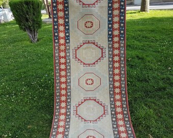 May include: A long, narrow rug with a cream background and red, blue, and white geometric patterns. The rug has a fringe on both ends.