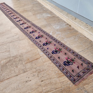 May include: A long, narrow runner rug featuring a dusty pink background with a repeating pattern of blue vases filled with pink and green flowers. The border displays a decorative floral motif in blue, green, and red, finished with fringe.
