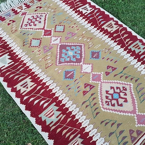 May include: A traditional style rug with a geometric pattern in shades of red, beige, blue, and white. The rug has a fringe border.