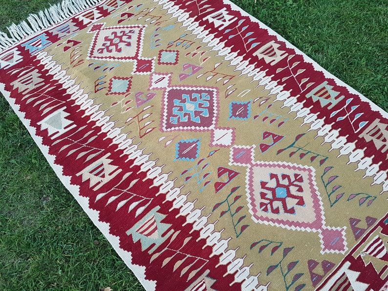 May include: A traditional style rug with a geometric pattern in shades of red, beige, blue, and white. The rug has a fringe border.