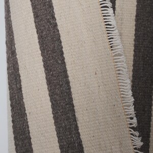 May include: A close-up of a woven rug with white and dark gray stripes. The rug has a fringe edge.