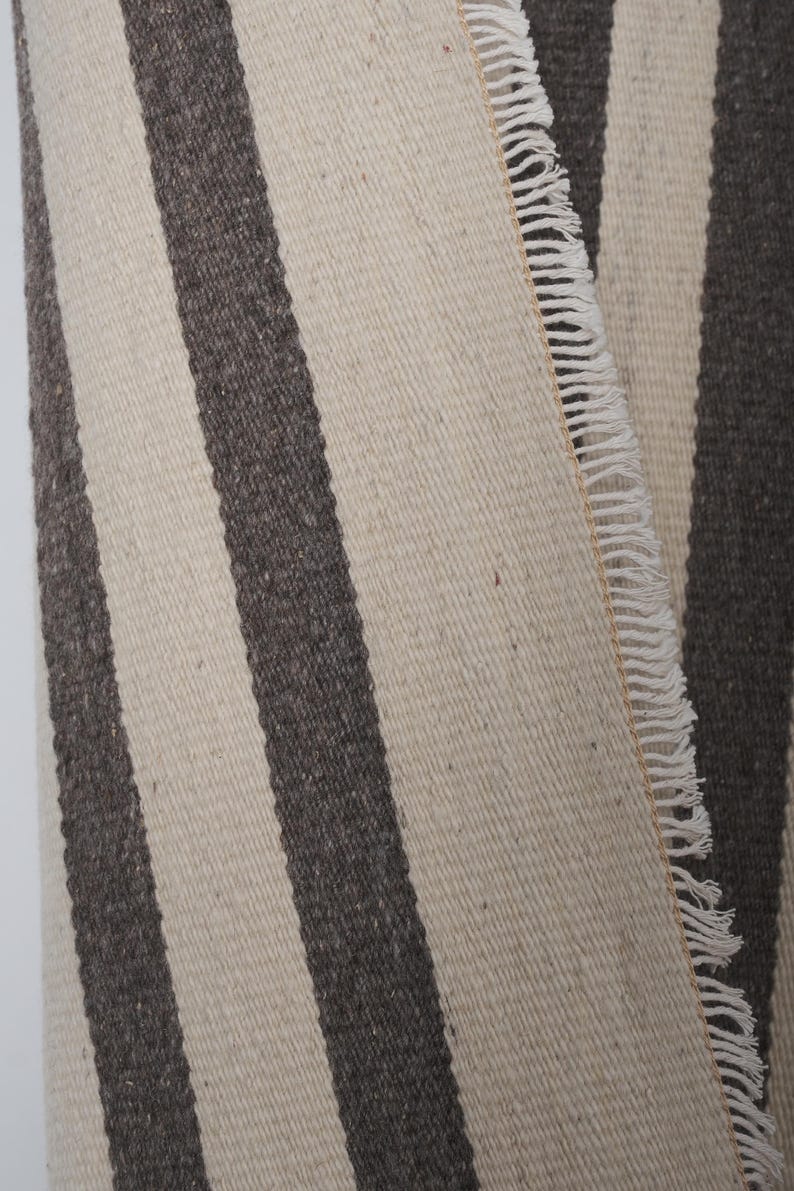 May include: A close-up of a woven rug with white and dark gray stripes. The rug has a fringe edge.