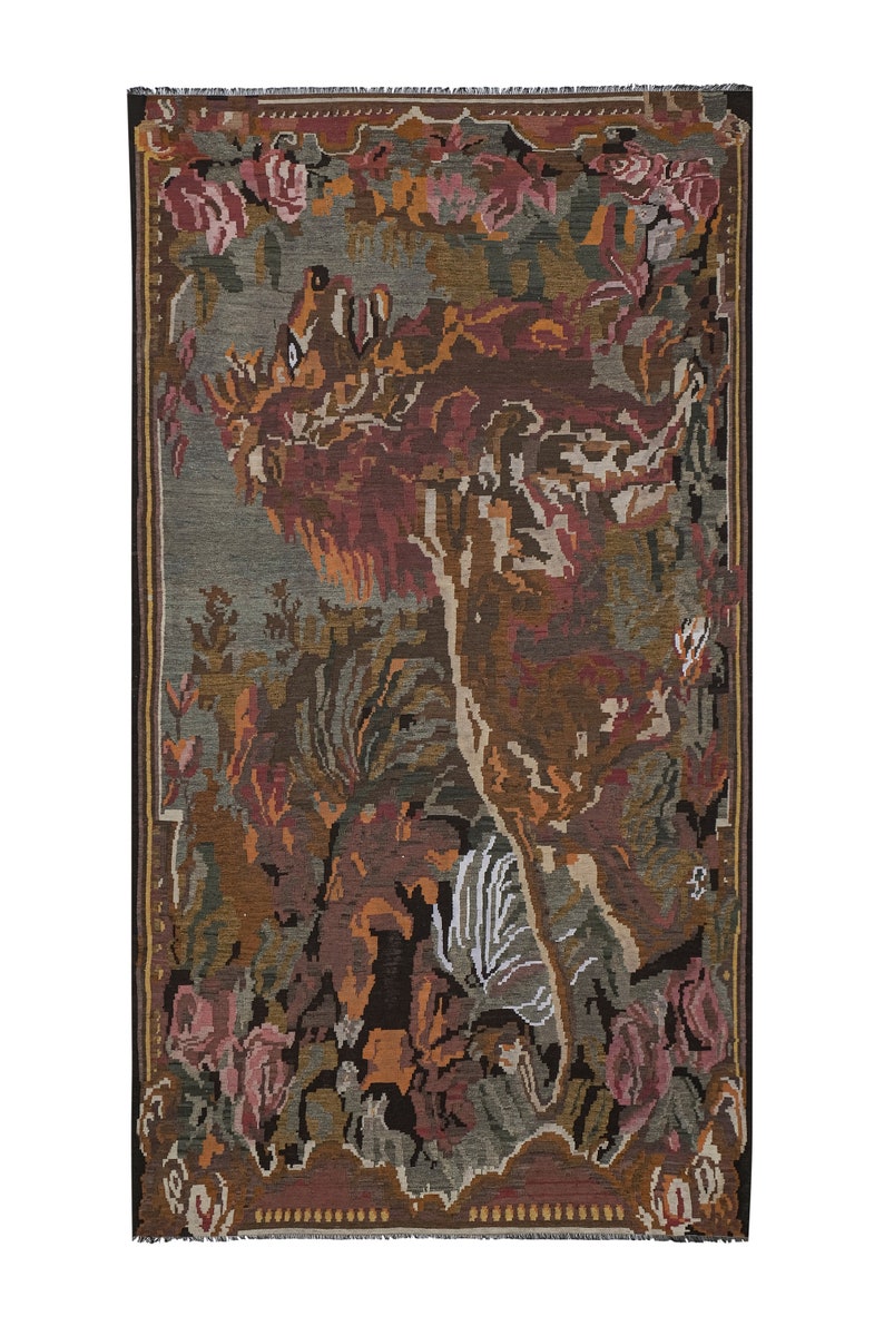 Op de afbeelding: Een rechthoekige wandtapijt met een gedetailleerd ontwerp met een leeuw, bloemen en bladeren in tinten bruin, rood, groen en grijs. Het wandtapijt heeft een donkere rand en een gefranjerde rand, geschikt voor wanddecoratie.
