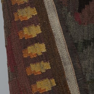 Op de afbeelding: Close-up van een geweven textiel met een geometrisch patroon. Het textiel heeft een kleurenpalet van bruin, rood en geel. De rand heeft een franje detail. Het textiel is waarschijnlijk een tapijt of wandkleed.