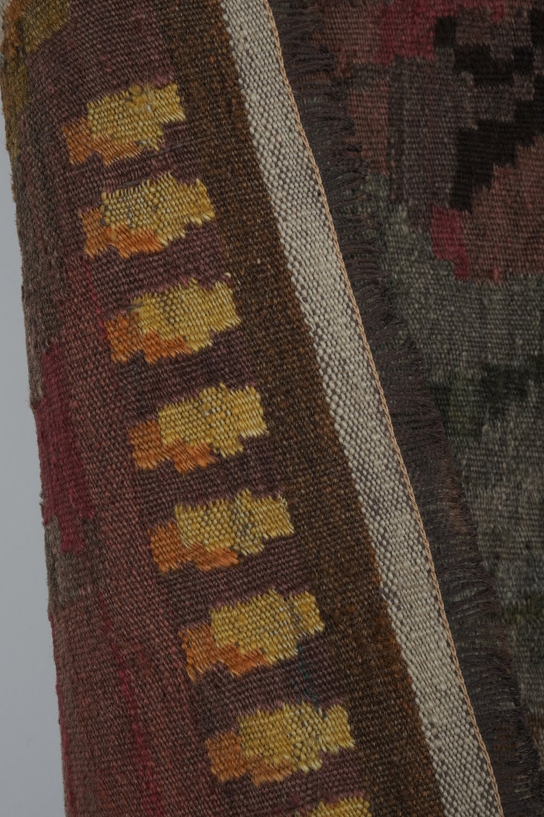 Op de afbeelding: Close-up van een geweven textiel met een geometrisch patroon. Het textiel heeft een kleurenpalet van bruin, rood en geel. De rand heeft een franje detail. Het textiel is waarschijnlijk een tapijt of wandkleed.