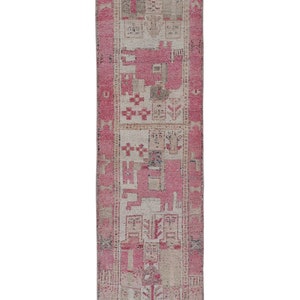 May include: A rectangular, vintage runner rug with a distressed appearance. The rug features a central design in shades of pink, beige, and gray, with a border in a darker pink. The pattern includes geometric and floral motifs.