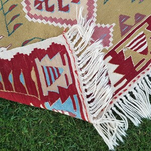 May include: A close-up of a colorful woven rug with a geometric pattern. The rug has a fringe border and is lying on a bed of green grass.