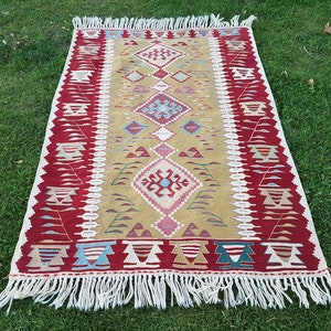 May include: A traditional Turkish kilim rug with a geometric pattern in shades of red, green, blue, and beige. The rug has a fringe border and is woven with wool.
