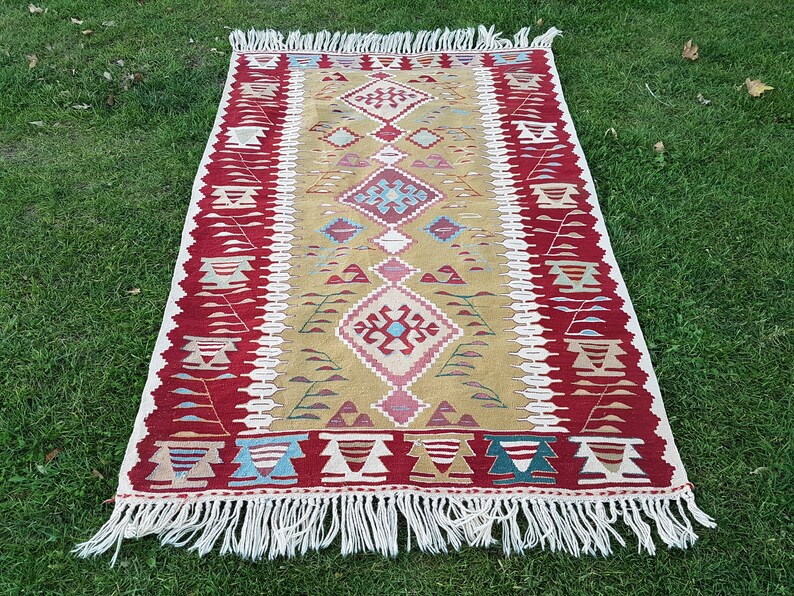 May include: A traditional Turkish kilim rug with a geometric pattern in shades of red, green, blue, and beige. The rug has a fringe border and is woven with wool.