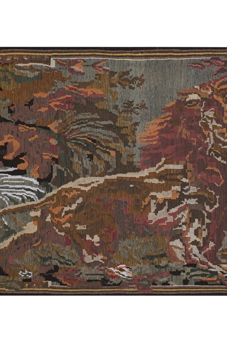 Op de afbeelding: Een geweven tapijt met een landschapssc&egrave;ne. Het tapijt heeft een donkerbruine rand en toont bomen, bladeren en een rivier in bruin-, rood- en groentinten. Het is een decoratief item.