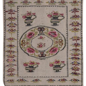 May include: Cream-colored, rectangular rug with a floral design. The rug features pink and green floral arrangements in vases and a central oval wreath of pink roses. The border has a repeating floral pattern and fringe.
