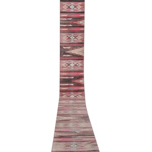 May include: A long, narrow rug with a geometric pattern in shades of pink, brown, and gray. The rug has a fringe on both ends.
