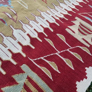 May include: Close-up of a red, white, and beige woven rug with a geometric pattern. The rug has a fringe border and is made of wool.