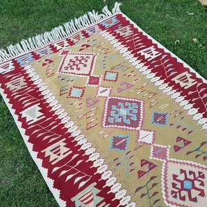 May include: A traditional Turkish kilim rug with a geometric pattern in shades of red, beige, and blue. The rug has a fringe border and is woven with wool.