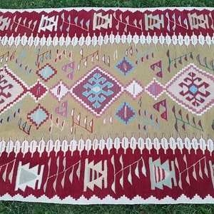 May include: A traditional Turkish kilim rug with a geometric pattern in shades of red, blue, white, and beige. The rug has a fringe border and is woven with a variety of textures.