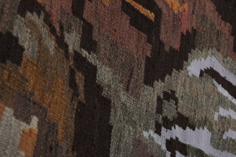 Op de afbeelding: Close-up van een geweven textiel met een geometrisch patroon. Het tapijt heeft een mix van kleuren, waaronder bruin, grijs, oranje en wit. Het ontwerp heeft een textuur, met zichtbare draden en een gedetailleerde weefsel.