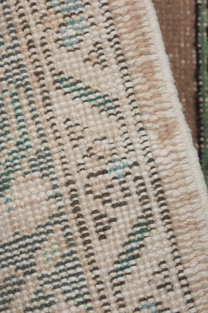 May include: Close-up of a beige rug with a woven pattern of black, green, and blue lines. The rug has a fringe edge.