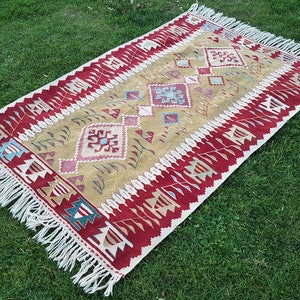 May include: A traditional Turkish kilim rug with a geometric pattern in shades of red, beige, and blue. The rug has a fringe border and is laid out on a green grassy surface.