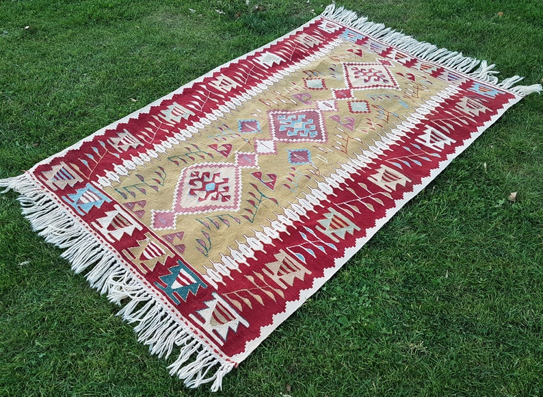 May include: A traditional Turkish kilim rug with a geometric pattern in shades of red, beige, and blue. The rug has a fringe border and is laid out on a green grassy surface.