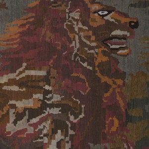 Op de afbeelding: Textielkunst die de kop en manen van een leeuw toont in tinten bruin, rood en oranje tegen een grijze achtergrond. Het kunstwerk heeft een gepixeleerd, geweven uiterlijk, wat duidt op een tapijt of vloerkleed.