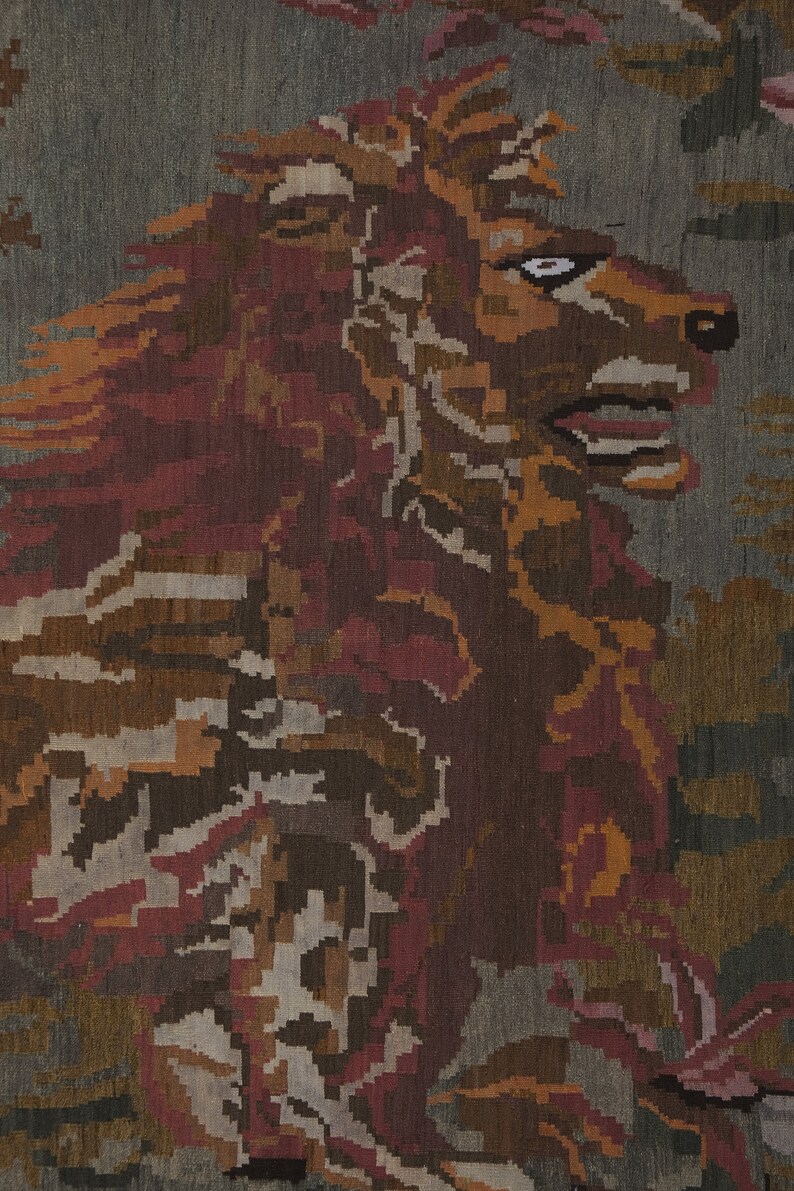 Op de afbeelding: Textielkunst die de kop en manen van een leeuw toont in tinten bruin, rood en oranje tegen een grijze achtergrond. Het kunstwerk heeft een gepixeleerd, geweven uiterlijk, wat duidt op een tapijt of vloerkleed.