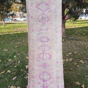 Könnte beinhalten: Ein langer, schmaler Teppich mit einem verblassten geometrischen Muster in Rosa und Weiß. Der Teppich ist aus Wolle und hat einen abgenutzten Look.