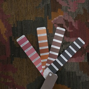 Op de afbeelding: Close-up van een patroontextiel met een kleurenpalet van bordeaux, oranje en grijs. Vier Pantone-kleurstalen waaieren uit en tonen tinten die het ontwerp van het textiel aanvullen. Het textiel heeft een geweven uiterlijk.
