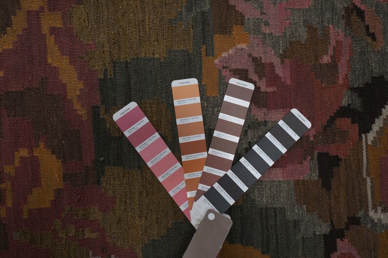 Op de afbeelding: Close-up van een patroontextiel met een kleurenpalet van bordeaux, oranje en grijs. Vier Pantone-kleurstalen waaieren uit en tonen tinten die het ontwerp van het textiel aanvullen. Het textiel heeft een geweven uiterlijk.