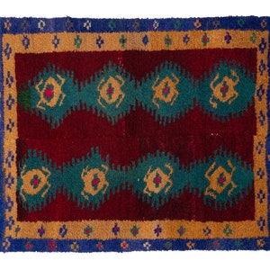 May include: A traditional rug with a red background and a geometric pattern in shades of green, blue, and yellow. The rug has a fringe border.