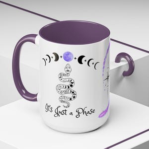 Peut inclure: Mug en céramique blanche avec intérieur, anse et accents violets. La tasse présente une illustration de serpent noir et blanc, des croissants de lune et le texte "It's Just a Phase". Un motif d'éclat d'étoile est sur le côté.