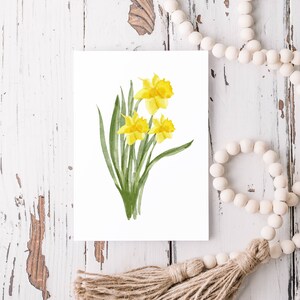 Puede incluir: Una pintura de acuarela de un ramo de narcisos amarillos con hojas verdes sobre un fondo blanco.
