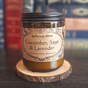May include: A glass jar candle with a black lid, labelled "Apothecary Blend Cucumber, Sage & Lavender." The candle sits on a wooden coaster. The label includes the website www.forageandfolkcollective.com. The candle has a warm, amber glow.