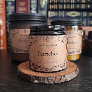 May include: Three apothecary jars with black lids and labels reading "Petrichor" and "Apothecary Blend". The jars are on a wooden slice, with a small brown bottle behind them. A shelf of books forms the background.