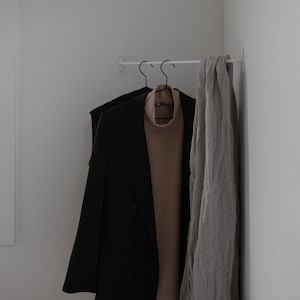 May include: A minimalist white wall-mounted clothing rack with black hangers. A black blazer, a tan turtleneck sweater, and a light gray scarf hang on the rack. The items are against a white wall, creating a clean, modern aesthetic.