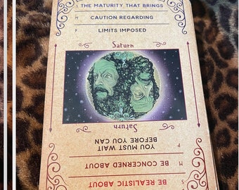 Puede incluir: Una carta del tarot con fondo marrón y bordes dorados ornamentados. La carta presenta una representación del planeta Saturno con dos caras, una verde y otra azul. El texto de la carta dice "Saturno" y "La madurez que trae." La carta también incluye el texto "Sé realista acerca de", "Preocúpate por", "Debes esperar antes de poder", "Límites impuestos", "Precaución con respecto a" y "La madurez que trae."