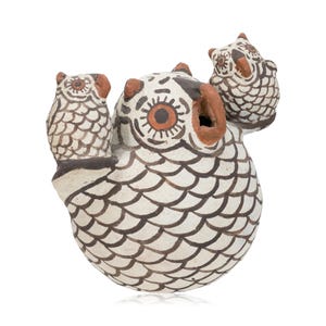 May include: A ceramic owl figurine with two smaller owls perched on its sides. The main owl is white with brown and black details, including scale-like patterns and large orange eyes. The figurine has a rounded body and a small opening.