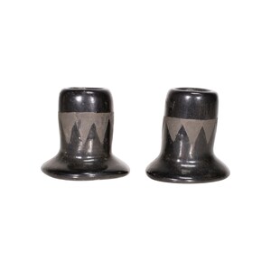 May include: Two black ceramic candle holders with a flared base and a cylindrical top. Each holder features a gray geometric design around the middle. The candle holders are displayed against a white background.