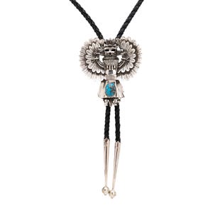 May include: A silver bolo tie featuring a detailed Native American figure with outstretched wings and a turquoise stone. The pendant is suspended from a black braided leather cord with silver tips and decorative beads. The design showcases intricate metalwork.