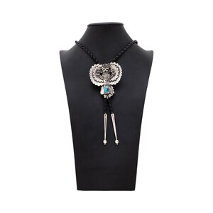 May include: Silver bolo tie with a detailed central pendant featuring a skull and feather design, accented with a turquoise-colored stone. The tie has a braided black cord and silver tips, displayed on a black mannequin.