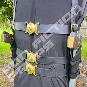Puede incluir: Un arnés de cuero negro con tres correas horizontales aseguradas con hebillas circulares doradas. El arnés incluye dos fundas laterales que sostienen réplicas de pistolas marrones y plateadas. El texto "TRAYWICK DESIGNS" es visible.
