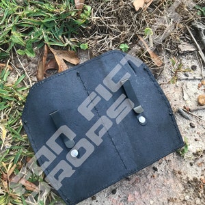 Op de afbeelding: Zwart leren holster met twee metalen clips. Het holster heeft twee vakken en is ontworpen om twee items te bevatten.