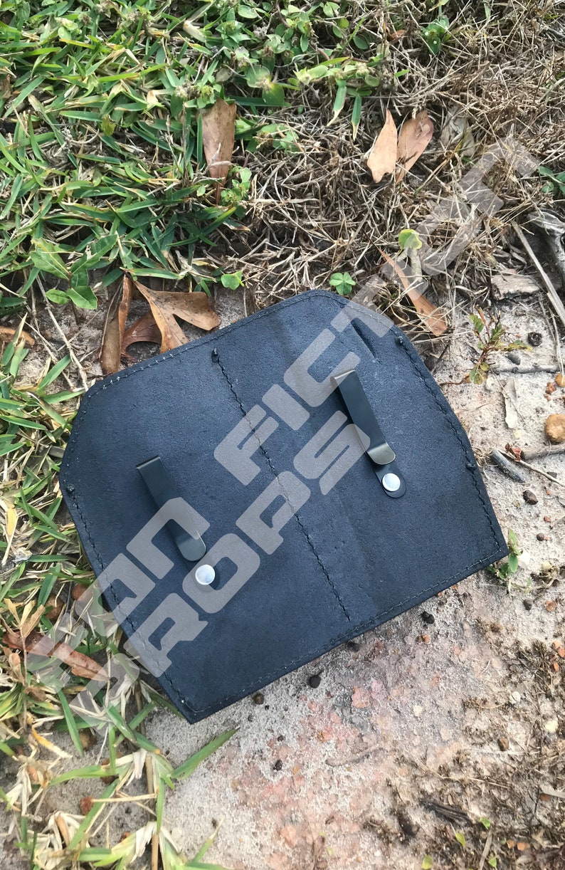 Op de afbeelding: Zwart leren holster met twee metalen clips. Het holster heeft twee vakken en is ontworpen om twee items te bevatten.