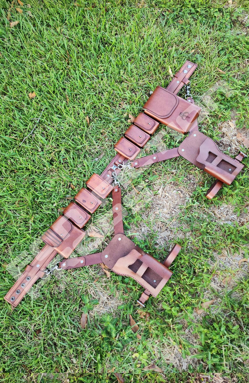 Puede incluir: Un cintur&oacute;n utilitario de cuero marr&oacute;n con m&uacute;ltiples bolsas y una funda, conectados por cadenas y correas de metal. El cintur&oacute;n est&aacute; dise&ntilde;ado con un patr&oacute;n de camuflaje y est&aacute; sobre hierba verde.