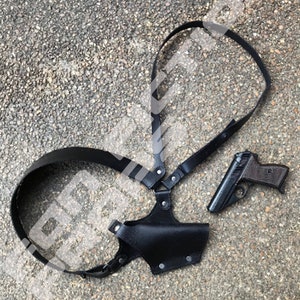 Könnte beinhalten: Ein schwarzes Leder-Schulterholster mit einer schwarzen Pistole. Das Holster hat verstellbare Riemen und einen Druckknopfverschluss.