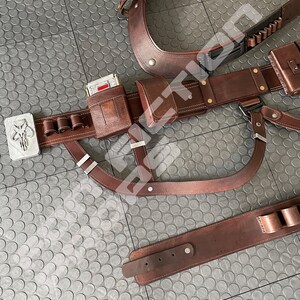 May include: A brown leather belt with multiple pouches and a silver buckle featuring a Mandalorian symbol. The belt is worn on a gray textured surface.