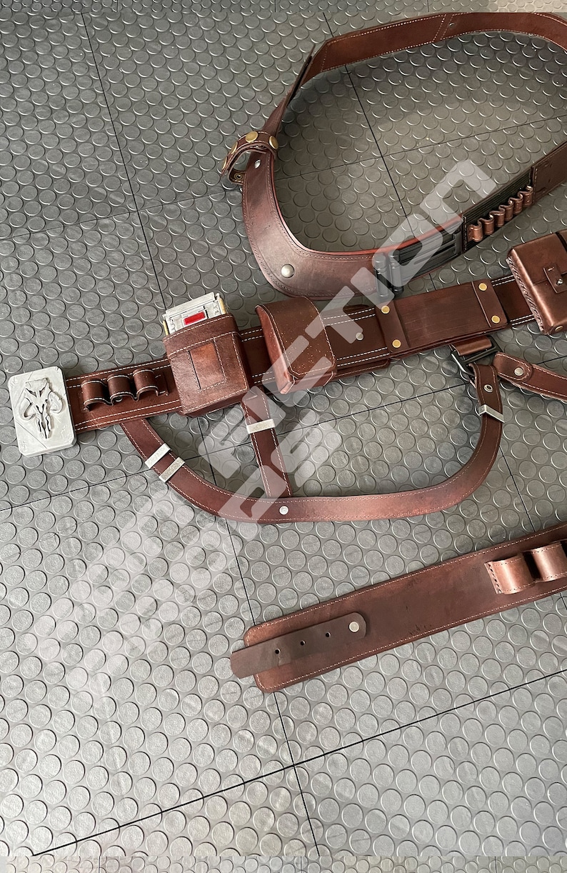 May include: A brown leather belt with multiple pouches and a silver buckle featuring a Mandalorian symbol. The belt is worn on a gray textured surface.