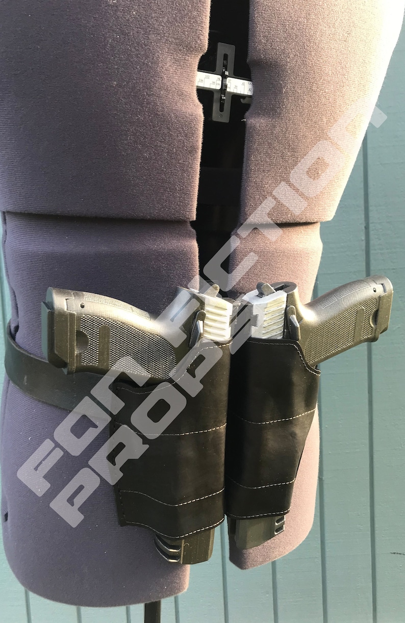 Op de afbeelding: Een zwart leren dubbel holster met twee zwarte handvuurwapens. De holsters zijn bevestigd aan een zwarte riem.