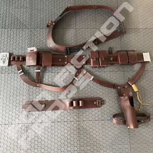 May include: A brown leather belt with multiple pouches and straps. The belt has a silver buckle and a silver skull emblem. The belt is laid out on a gray textured surface.